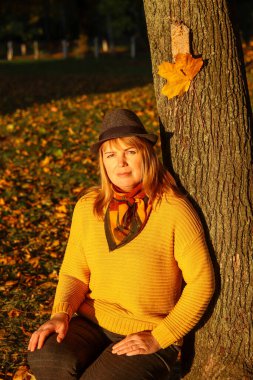 Renkli turuncu sonbahar parkındaki güzel sarışın kadının portresi. Şapkalı ve ağaç gövdesinin yanında örgü ören şık bir kadın. Merhaba sonbahar konsepti. Bulanık sarı akçaağaç yaprağı. Aşk doğası.