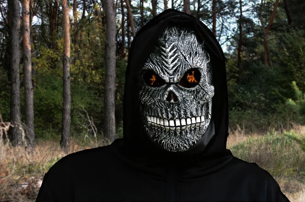 Close-up portrait of grim reaper. Man in death mask with fire flame in eyes on dark autumn forest pine tree background. Carnival costume, creepy teeth. Halloween holiday concept. Dark horror