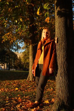 Sonbahar parkında beyaz saçlı genç bir kadının tam boy portresi. Sarı kazaklı, kahverengi paltolu ve parkta duran parlak stil sahibi bir kadın. Mutlu sonbahar konsepti. Güneşli hava, gün batımı.