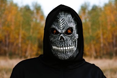 Cadılar bayramı balkabağı kafasını tutan ölüm meleği. Ölüm maskeli, sarı sonbahar ormanı arka planında gözleri alev alev yanan bir adam. Cadılar Bayramı konsepti. Karanlık korku