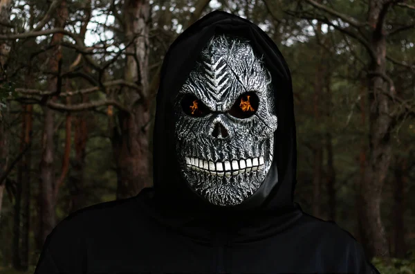 Close-up portrait of grim reaper. Man in death mask with fire flame in eyes on dark forest autumn background. Carnival costume, creepy teeth. Halloween holiday concept. Dark horror