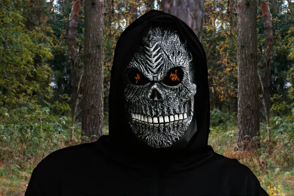 Close-up portrait of grim reaper. Man in death mask with fire flame in eyes on nature forest tree trunk background. Carnival costume, creepy teeth. Halloween holiday concept. Dark horror.
