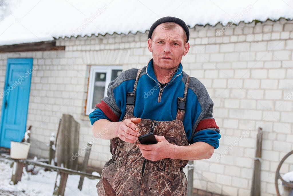 Russian young man standing outside on winter countryside background. Construction worker holding phone. Caucasian laborer man 40 years old, portrait. Poverty concept. Unhappy sad people.