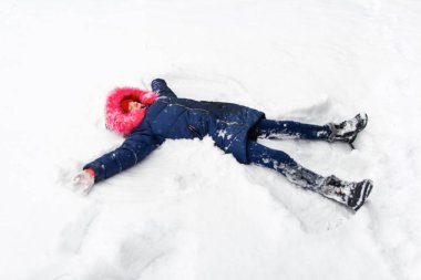 Pembe kapüşonlu mutlu genç kız kar meleği yaparken arka planda kar üzerinde yatıyor. Dışarıda güzel bir çocuk, soğuk bir hava. Kafkasyalı çocuk. İyi tatiller. Kış keyifli.