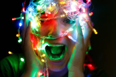 Kadın panik içinde başını sıkıca tutuyor. Stresli Noel Kızı Çığlığı. Genç insan kış tatilinde renkli ışıklarla eğleniyor. Renkli Noel ışıklarına sarılmış bir kadın.