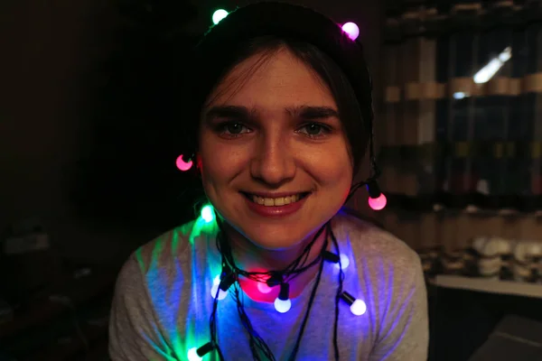 Julnärbild leende porträtt. En lustig ung kvinna som firar jul. Ung person som har roligt på vintern semester med färgglada ljus. Kvinna insvept i färgglada julbelysning — Stockfoto