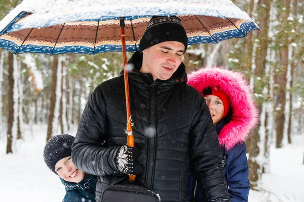 Genç bir adam ve komik çocuklar kırsal kesimdeki karlı arka planda şemsiye tutuyorlar. Mutlu aile, soğuk hava. Yaşam tarzı. Tatiller. Karlı kış, ormanda oynamak. Ağabey. Aptal!