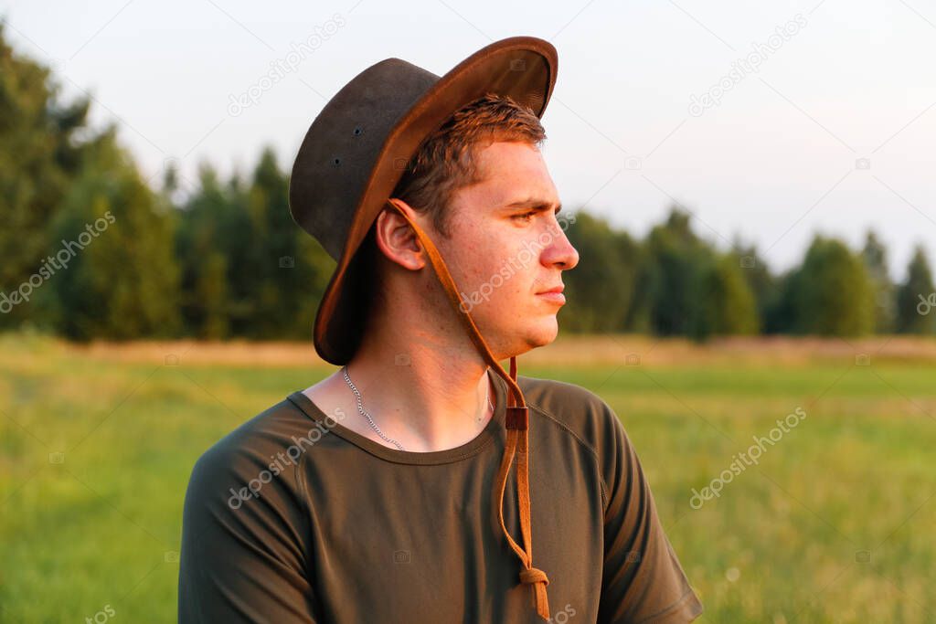 Young man farmer in cowboy hat at agricultural field on sunset with sun flare. Portrait of millennial man with hat, standing on grass background, outdoors in meadow. Farming, harvest concept.