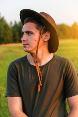 Young man serious farmer in cowboy hat at agricultural field on sunset with sun flare. Profile portrait of man standing on nature background, outdoors. Rancher on farmland. Happy summer. Close-up.