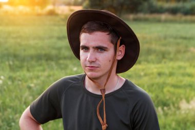 Young man farmer in cowboy hat at agricultural field on sunset with sun flare. Closeup portrait of millennial man with hat, standing on nature background, outdoors. Countryman, rancher, cowboy.