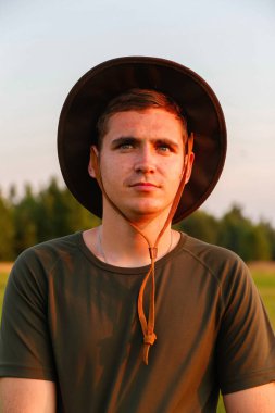 Young man serious farmer in cowboy hat at agricultural field on sunset with sun flare. Portrait of millennial man standing on nature background, outdoors. Rancher on farmland. Happy summer. Close-up.