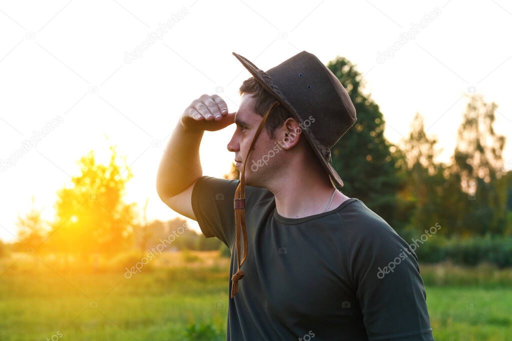 Young man farmer in cowboy hat at agricultural field on sunset with sun flare. Closeup portrait of millennial man with hat, standing on nature background, outdoors. Rancher look in to distance.