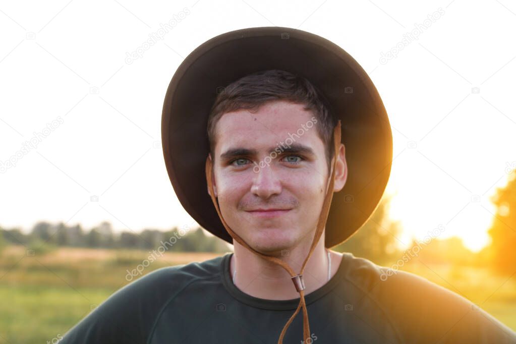 Young man farmer in cowboy hat at agricultural field on sunset with sun flare. Closeup portrait of millennial man with hat, standing on nature background, outdoors. Rancher.