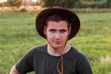Young man farmer in cowboy hat at agricultural field on sunset with sun flare. Closeup portrait of millennial man with hat, standing on nature background, outdoors. Countryman, rancher on farmland.