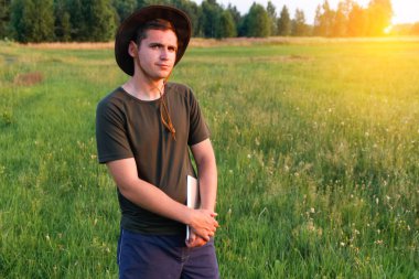 Young man farmer in cowboy hat at agricultural field on sunset holding tablet. Male on nature background, outdoors in meadow. Agriculture concept. Digital farmland. Smart agronomist. Copy space.