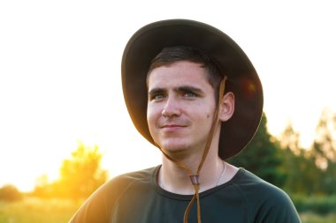 Young man smiling farmer in cowboy hat at agricultural field on sunset with sun flare. Closeup portrait of millennial man standing on nature background, outdoors. Countryman on farmland. Happy summer.
