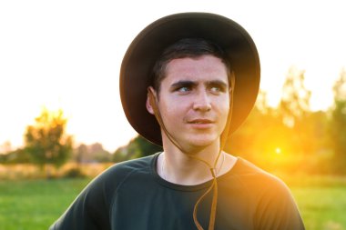 Young man smiling farmer in cowboy hat at agricultural field on sunset with sun flare. Closeup portrait of millennial man standing on nature background, outdoors. Rancher on farmland. Happy summer.