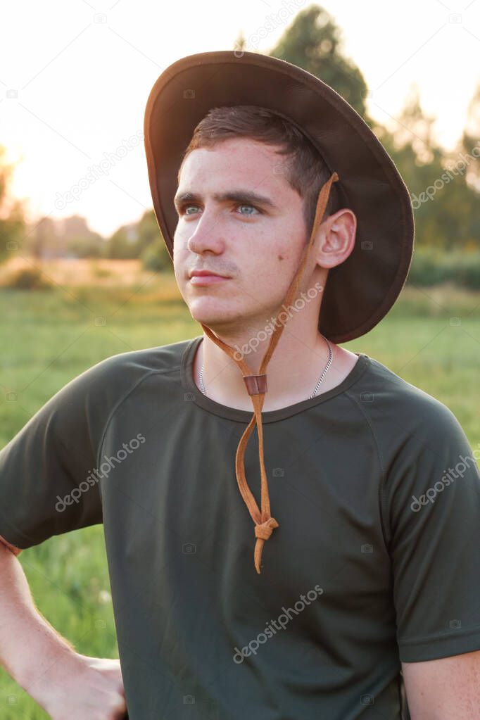 Young man farmer in cowboy hat at agricultural field on sunset with sun flare. Closeup portrait of millennial man with hat, standing on nature background, outdoors. Countryman, rancher.