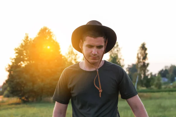 Jeune homme agriculteur en chapeau de cow-boy au champ agricole au coucher du soleil avec éruption de soleil. Gros portrait de l'homme millénaire avec chapeau, debout sur le fond de la nature, à l'extérieur. Rancher, ouest sauvage — Photo