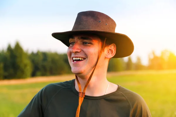 Молодой человек улыбается фермеру в ковбойской шляпе на сельскохозяйственном поле на закате с солнечной вспышкой. Откровенный портрет человека тысячелетия в шляпе, стоящего на природе, на открытом воздухе. Сельский житель на ферме — стоковое фото