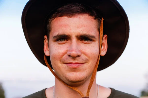 Молодой человек улыбается фермеру в ковбойской шляпе на сельскохозяйственном поле против голубого неба. Портрет человека тысячелетия, стоящего на природе, на открытом воздухе. Ранчо на ферме. Счастливого лета. Крупный план — стоковое фото