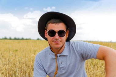 Güneş gözlüklü adam. Arka planda mavi gökyüzü olan altın buğday tarlasında duran çiftçi portresi. Tarlada buğday hasadını inceleyen güneş gözlüğü ve kovboy şapkası takan genç adam. Yulaf endüstrisi. Yakın çekim..