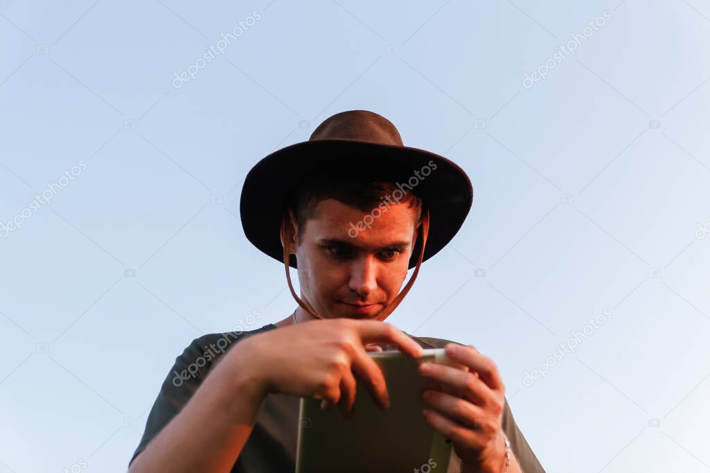 Young man affected farmer in cowboy hat at agricultural field holding tablet. Male on blue sky background, outdoors in meadow. Agriculture concept. Digital farmland. Smart agronomist. Analysis.