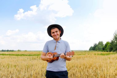 Şarkıcı. Genç, güzel beyaz bir adam dışarıda ukulele çalıyor. Yakışıklı hippi, doğada gitar çalıyormuş. Kovboy şapkalı bir erkek gülümseyerek gitar çalıyor..