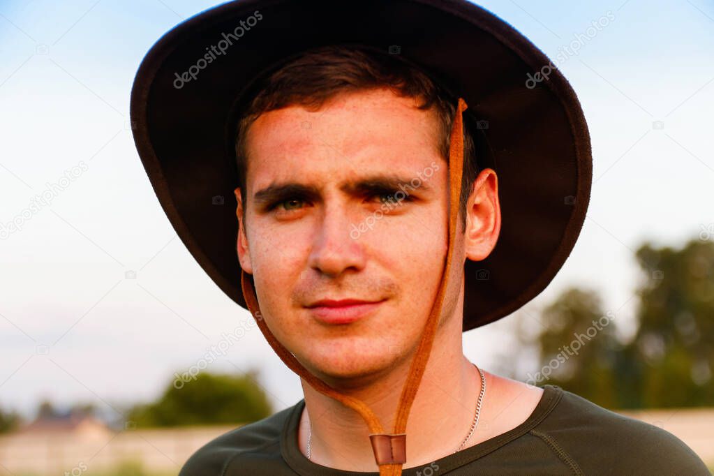 Young man smiling farmer in cowboy hat at agricultural field against blue sky. Portrait of millennial man standing on nature background, outdoors. Rancher on farmland. Happy summer. Close-up.
