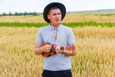 Hawaii gitarı. Genç, güzel beyaz bir adam dışarıda ukulele çalıyor. Yakışıklı hippi, doğada gitar çalıyormuş. Kovboy şapkalı bir erkek gülümseyerek gitar çalıyor..