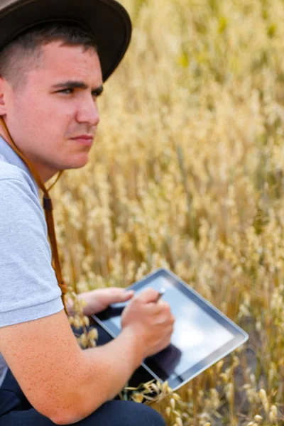Un joven granjero. Retrato del agricultor sentado en el campo de trigo dorado y desplazándose en la tableta. Joven con sombrero de vaquero en el campo examinando la cosecha de trigo. Industria del grano de avena. Borrosa. La avena planta. Copiar espacio —  Fotos de Stock