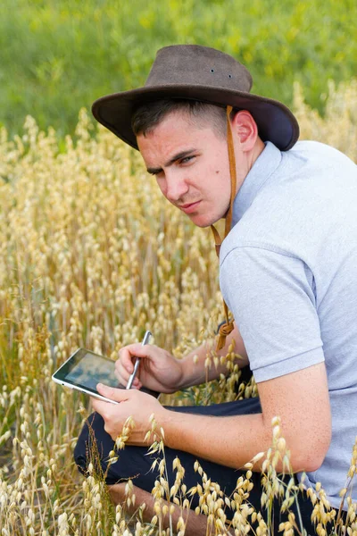 디지털 농장. 금밀 밭에 앉아 타블렛 위를 걷는 농부의 모습. 카우보이 모자를 쓴 젊은이 가 밭에서 밀 작물을 조사하고 있습니다. 귀리 곡물 산업. 필드. 귀리 식물 — 스톡 사진