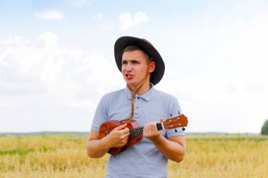 Yaz eğlencesi. Genç, güzel beyaz bir adam dışarıda ukulele çalıyor. Yakışıklı hippi, doğada gitar çalıyormuş. Kovboy şapkalı bir erkek gülümseyerek gitar çalıyor.