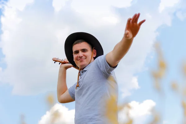 Músico de emoción. Joven hermoso hombre caucásico jugando ukelele fuera. Guapo chico hipster toca la guitarra ukelele en el fondo de la naturaleza. Macho en un sombrero vaquero sonríe y toca la guitarra — Foto de Stock