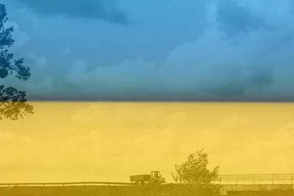 Ukrayna savaşı. Ukrayna bayrağı. Yeşil römorklu kırmızı bir traktör kırsal kesimde rüzgarlı bir havada köprüye çarpıyor. Çimleri, ağaçları, bulutları görebilirsin.