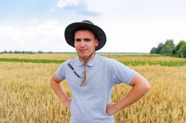 Komik hasatçı. Arka planda mavi gökyüzü olan altın buğday tarlasında duran çiftçi portresi. Tarlada buğday tarlasında güneş gözlüğü takan ve kovboy şapkası takan genç bir adam. Yulaf tohumu.