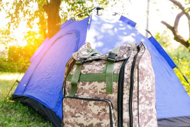 Yeşil doğa arka planında duran Defocus mavi açık turist çadırı. Ordu sırt çantası. Turizm konsepti. Ormanda yaz tatili, kamp. Vahşi doğa. Yürüyüş. - Yakın çekim. Güneş parlaması. Odaklanamıyorum.