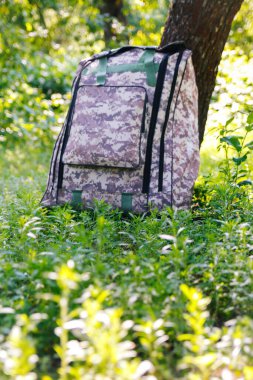 Askeri sırt çantasından çık. Ağacın yanındaki yeşil çimlerin üzerinde ordu çantası. İngiliz ordusunun sırt çantası ve sırt çantasında askeri kamuflaj ağı malzemesi. Dikey. Odaklanamıyorum