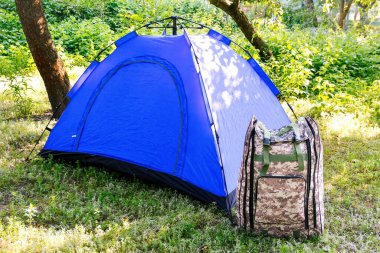 Yeşil doğa arka planında duran Defocus mavi açık turist çadırı. Ordu sırt çantası. Turizm konsepti. Ormanda yaz tatili, kamp. Yaşam tarzı. Odaklanamıyorum.