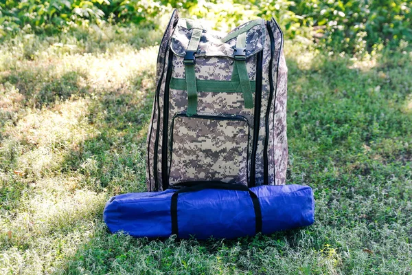 Defocus military backpack and blue tent or sleeping bag. Army bag on green grass background near tree. Military camouflage army rucksack. Vertical. Tourist summer hiker. Trip concept. Out of focus.