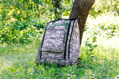 Askeri sırt çantasından çık. Ağacın yanındaki yeşil çimlerin üzerinde ordu çantası. İngiliz ordusunun sırt çantası ve sırt çantasında askeri kamuflaj ağı malzemesi. Turist yaz yürüyüşçüsü. Seyahat konsepti. Odaklanamıyorum.