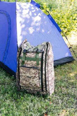 Yeşil doğa arka planında duran Defocus mavi açık turist çadırı. Ordu sırt çantası. Turizm konsepti. Ormanda yaz tatili, kamp. Yaşam tarzı. Yürüyüş. - Yakın çekim. Odaklanamıyorum.