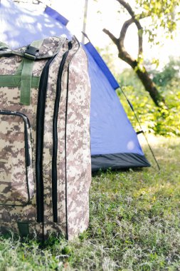 Yeşil doğa arka planında duran Defocus mavi açık turist çadırı. Ordu sırt çantası. Turizm konsepti. Ormanda yaz tatili, kamp. Vahşi doğa. Yürüyüş. - Yakın çekim. Dikey. Odaklanamıyorum.