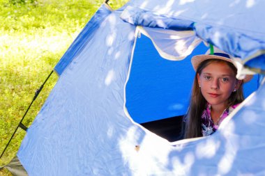 Defocus teen girl looks out of the tent window in camping. Green nature background. Forest or garden. Happy, rest, travel. Sunny. Blurred. Little girl in camp. Out of focus.