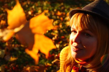 Defocus, sonbahar parkında gülümseyen beyaz, sarışın bir kadının yakın plan portresi. Turuncu arka planda şapkalı şık bir kadın. Mutlu sonbahar rüya konsepti. Bulanık sarı akçaağaç yaprağı. Odaklanamıyorum.