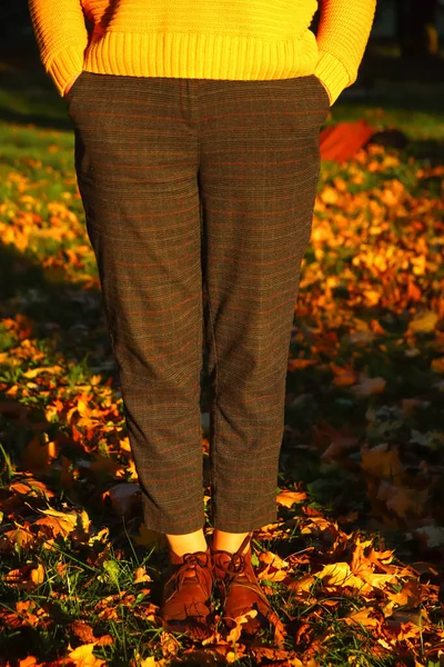 Defocus female legs in capri pants and brogues shoes on green grass with bright leaves. Bright stylish woman in orange coat walking in october park. Out of focus.