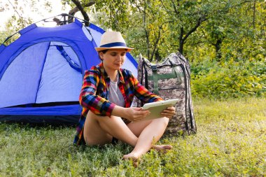 Defocus young woman working on tablet near camping tent outdoors surrounded by beautiful nature. Freelance, sabbatical, mental health. Okay gesture. Remote work. Summer tourist. Out of focus.