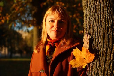 Defocus portrait of pretty Caucasian blonde woman in colorful dark maple autumn park. Bright stylish woman in coat near tree trunk. Hello fall concept. Blurred yellow maple leaf. Out of focus.