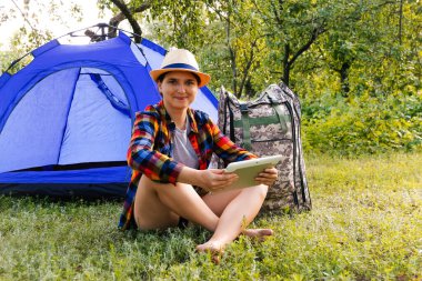 Defocus young woman working on tablet near camping tent outdoors surrounded by beautiful nature. Freelance, sabbatical, mental health. Okay gesture. Remote work. Summer camp. Burnout. Out of focus.