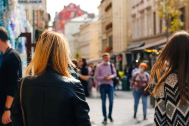 Defocus two stylish blonde and white women walking outdoors in autumn city street at sunset time wearing black jacket. View from the back. Friendship and travel. Out of focus.